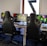 A row of gaming stations equipped with large monitors, gaming chairs, keyboards, and headphones. The setup is organized and ready for gaming, featuring a colorful screen display. The room has a modern and tech-savvy ambiance.