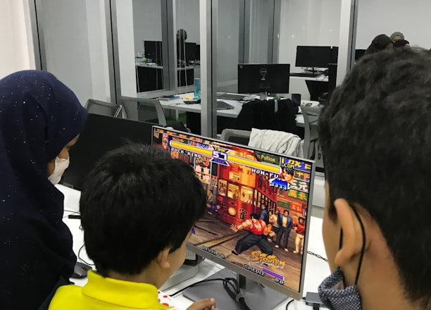 A group of children is gathered around a desktop computer, engaged in playing a colorful, action-packed arcade game displayed on the monitor. The setting appears to be a modern, well-lit office or gaming space with large glass windows, showing multiple computer workstations in the background.